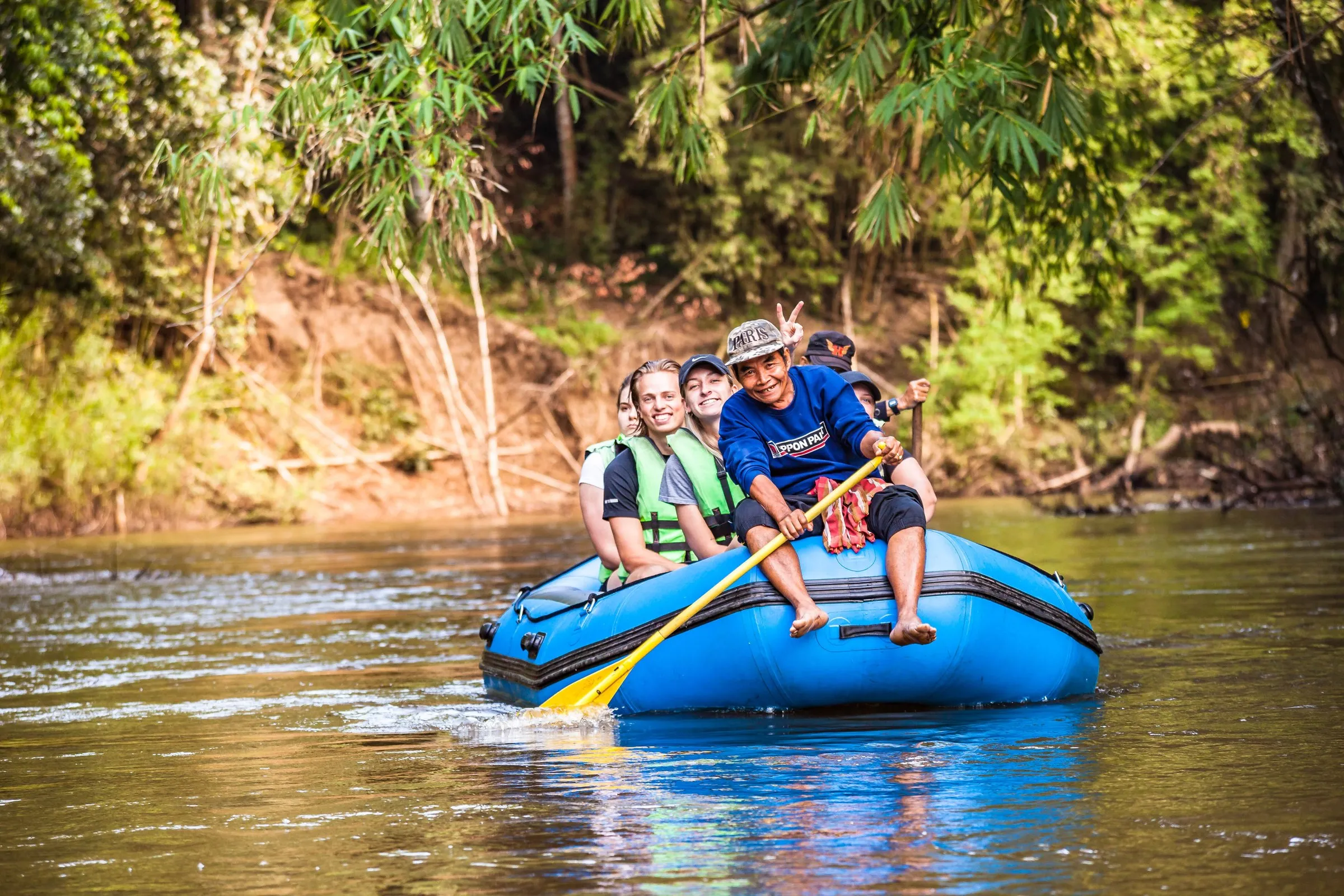 A smiling local guide paddling a blue raft full of travellers down the river.