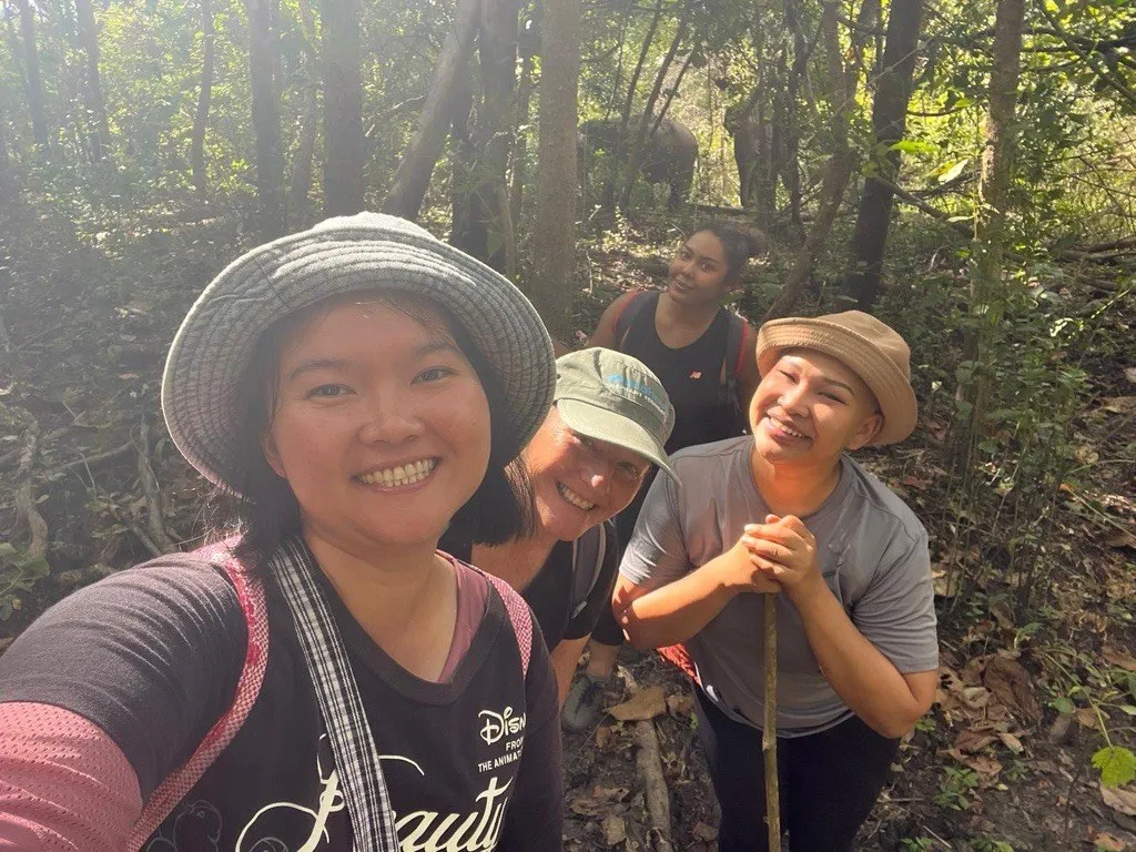 Smiling group selfie on the forest trail, elephants between the trees behind.